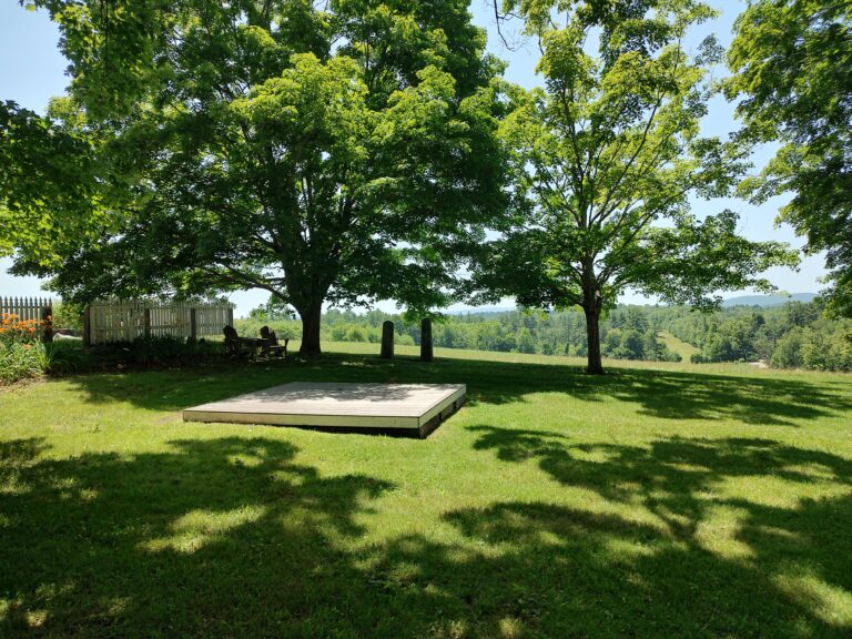 Sunlit park scene with two large trees over a grassy lawn, a wooden platform deck on the left, and a fence in the background.