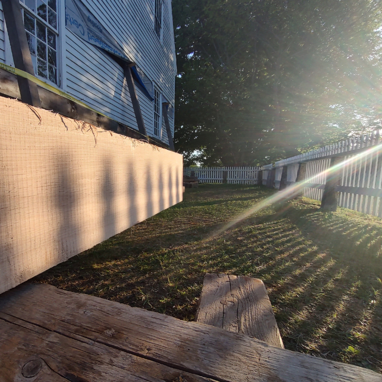 Construction site by a blue house with plywood barrier along the foundation, wooden deck planks in foreground, and a white picket fence in the background with sun rays near dusk/light.