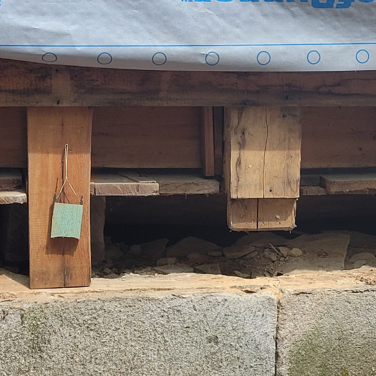 Temporary wooden shoring under a raised platform with a teal sponge hanging from a post under a tarp-covered area at a construction site