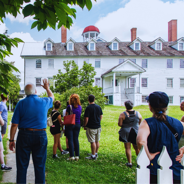 Tour group gathered on a lawn, facing a white historic mansion with dormers and a red cupola.