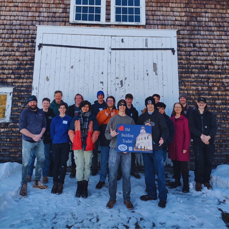 Group of about 15 people posing outdoors in front of a weathered wooden building with large white doors, snow on the ground; two hold a sign that reads 'Old Building Trades'.